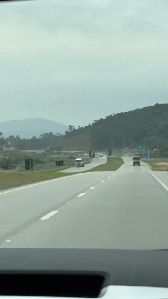 Rodovia vista do para-brisa, caminhões à frente e morros verdes ao fundo