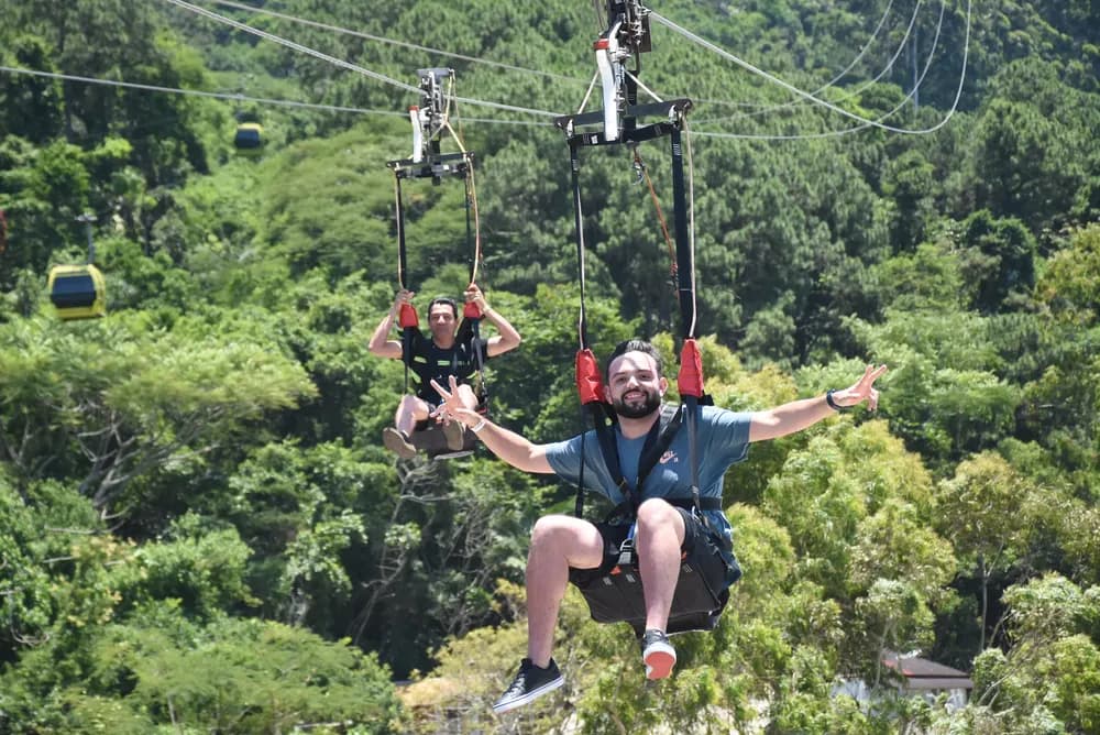 Duas pessoas em tirolesa sobre a mata, bondinho amarelo ao fundo