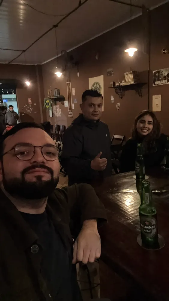 Selfie de três amigos num bar, garrafa de cerveja no balcão de madeira