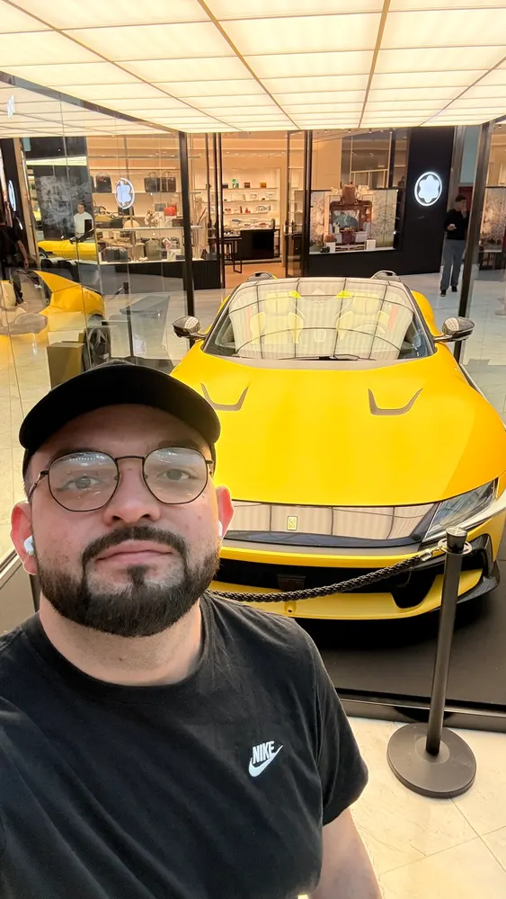 Selfie de boné e óculos diante de um carro esportivo amarelo exposto