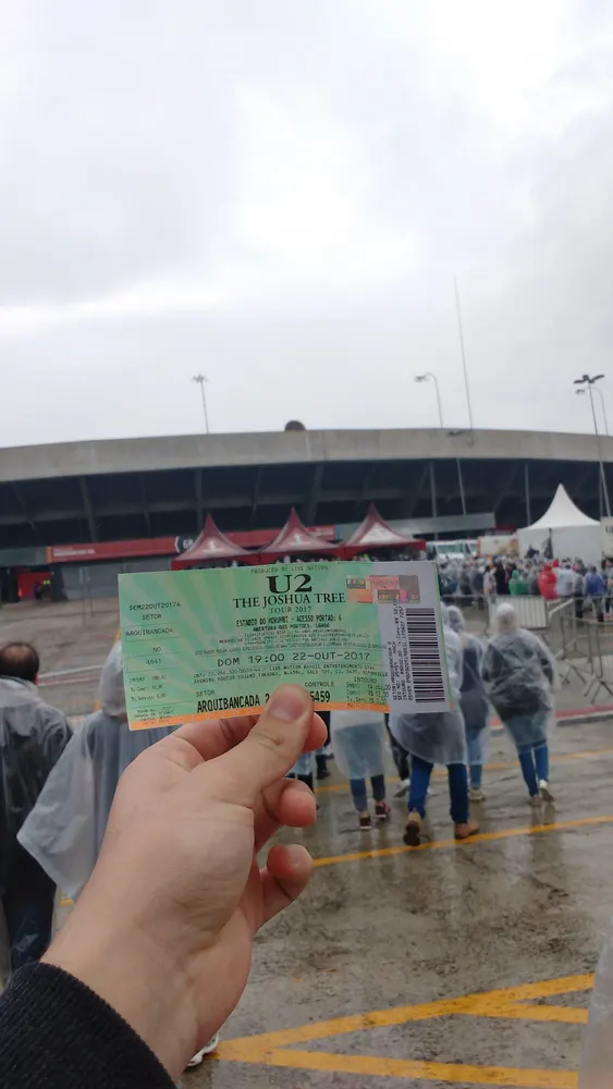 Mão segurando ingresso do U2 em frente a um estádio, dia chuvoso