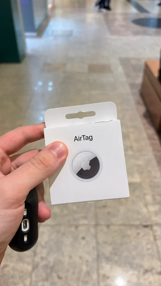 Caixa de um AirTag e um estojo preto na mão, num piso claro e polido