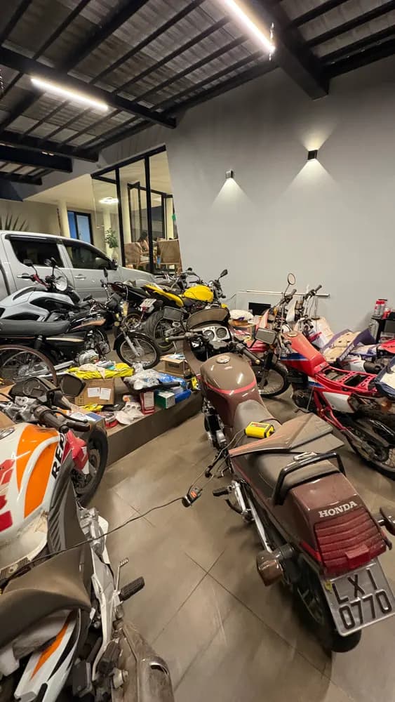 Garagem com várias motos, peças espalhadas e um carro ao fundo