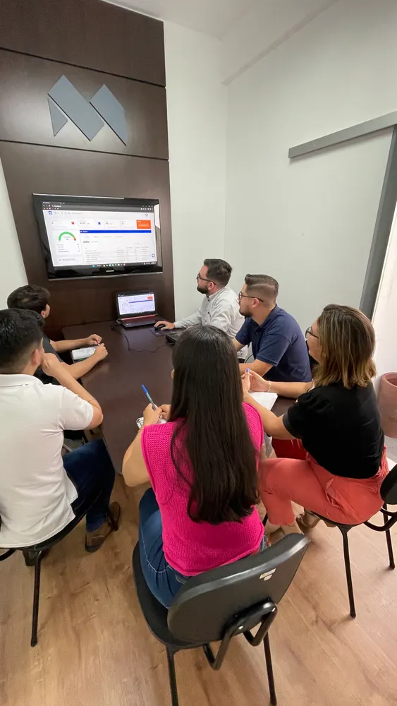 Reunião em sala pequena, grupo olha um painel de dados na TV