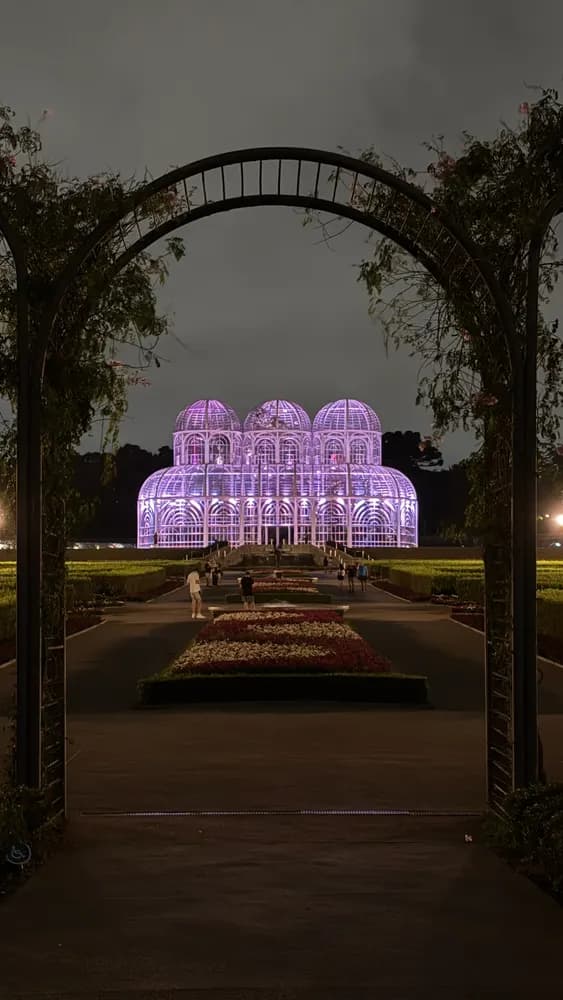 Estufa de vidro iluminada de roxo à noite, vista por um arco de plantas