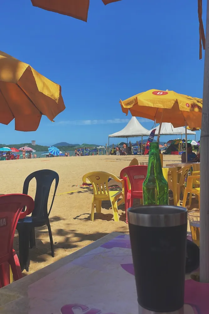 Mesa na praia com copo e garrafa verde, cadeiras coloridas e guarda-sóis