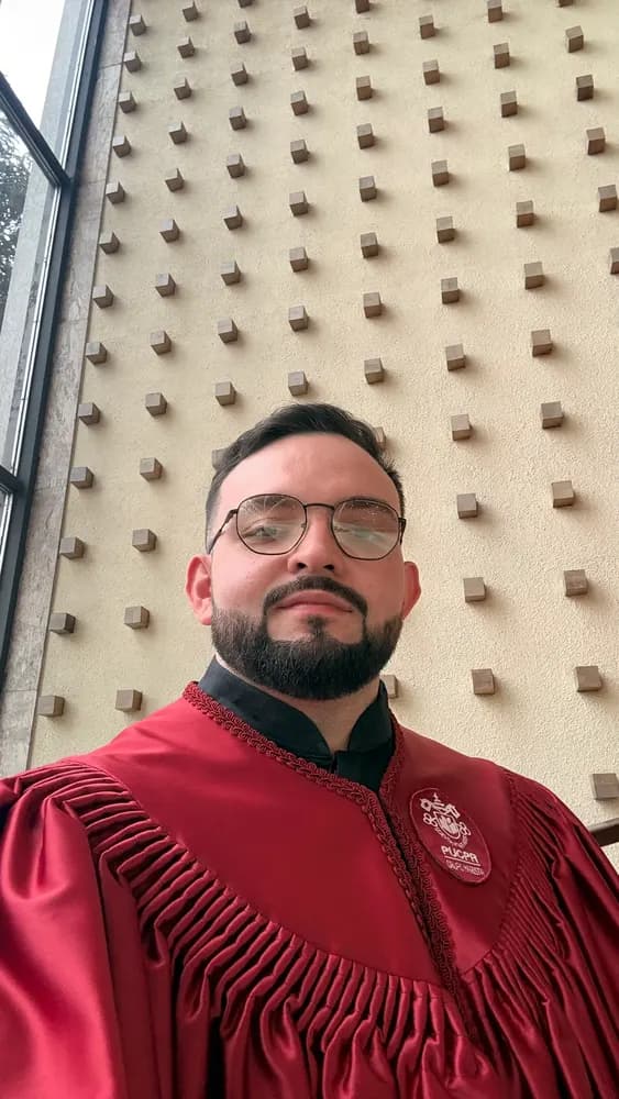 Homem de beca vermelha em selfie, parede bege com cubos em relevo