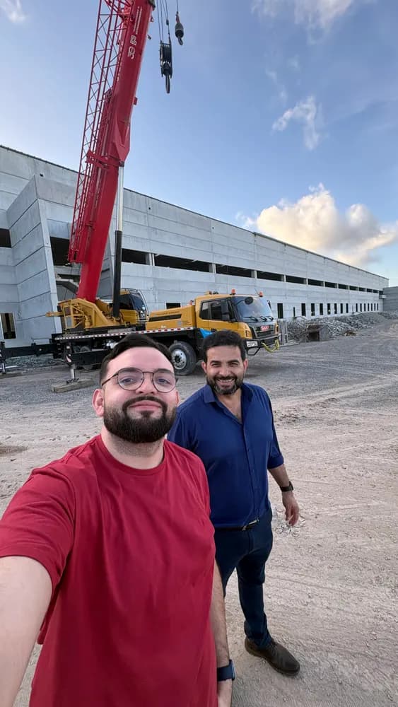 Duas pessoas sorriem em obra com guindaste vermelho e galpão de concreto