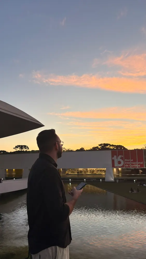 Homem de óculos diante de um espelho d'água, céu alaranjado no pôr do sol