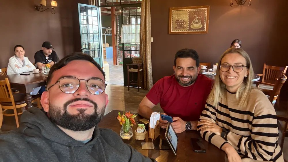 Selfie de três amigos à mesa de uma cafeteria de paredes marrons