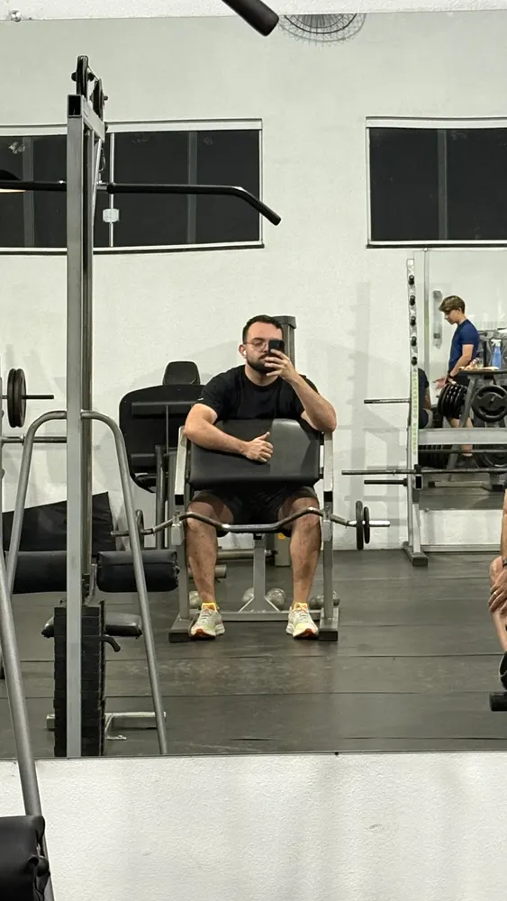 Homem com celular sentado em aparelho de musculação, refletido no espelho