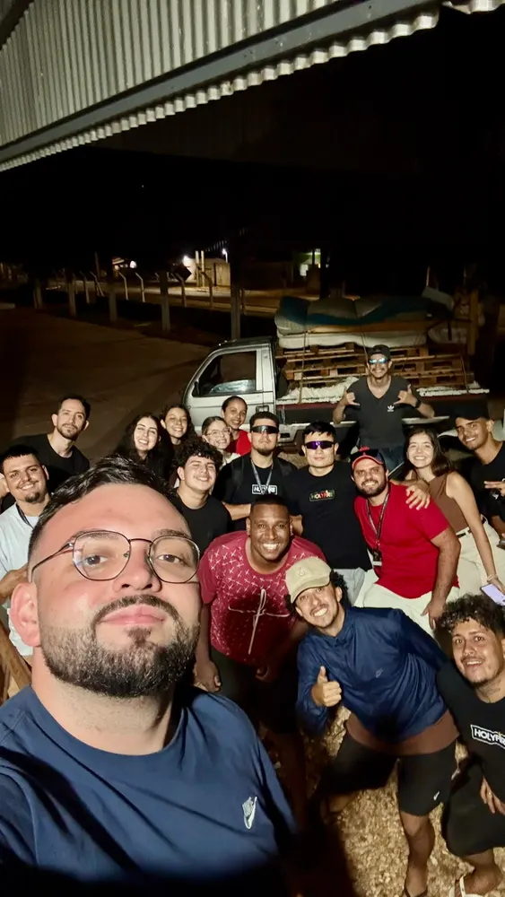Selfie à noite com um grupo grande de jovens, galpão e caminhão ao fundo