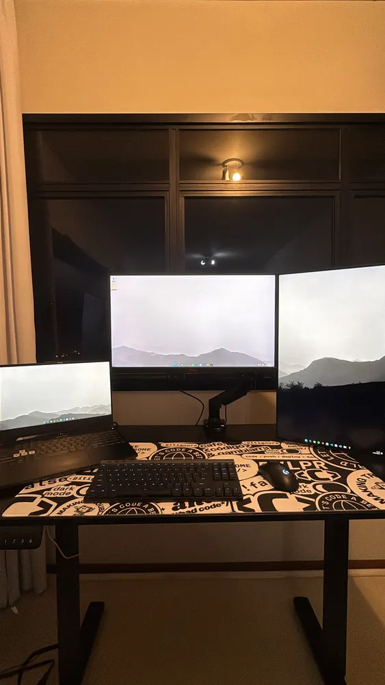 Mesa com notebook e dois monitores ligados, teclado e mouse, luz baixa