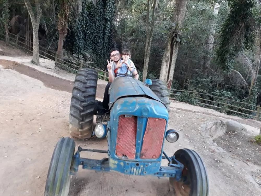 Homem e criança sobre um trator azul antigo em estrada de terra na mata