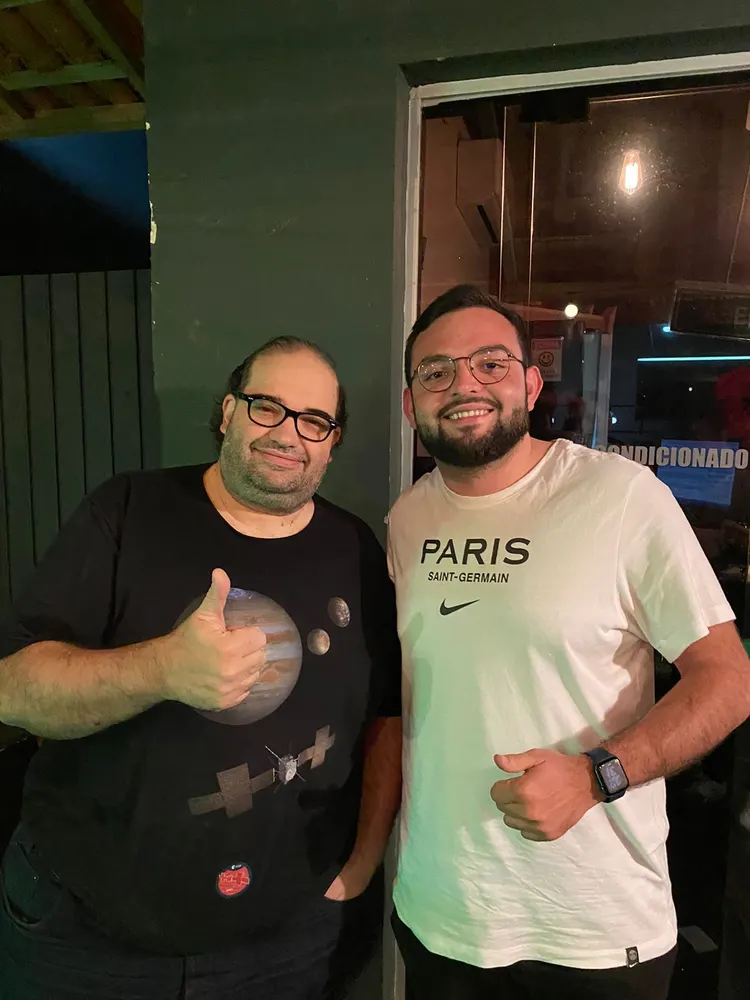 Dois homens de óculos posam à noite, um deles faz sinal de positivo